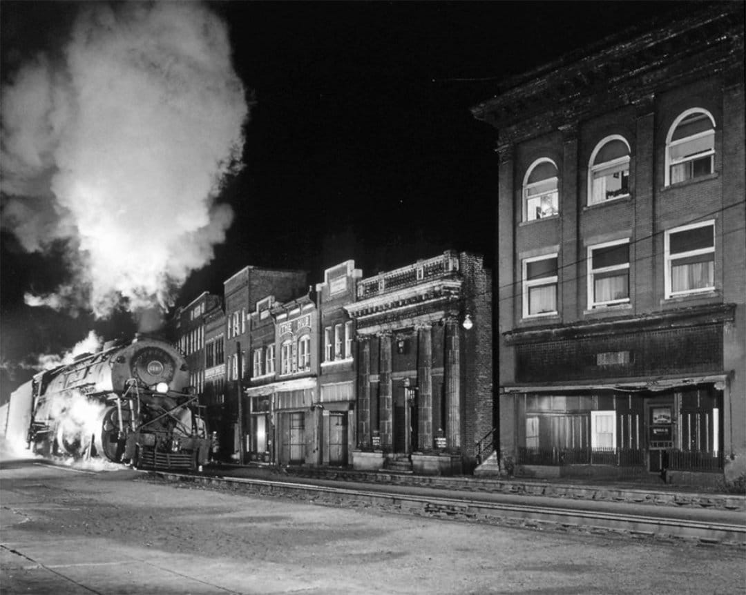 a row of three and two story city buildings with a steam engine approaching in front of them